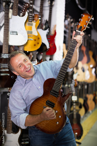 happy adult male is playing on electric guitar and satisfied it in music store.