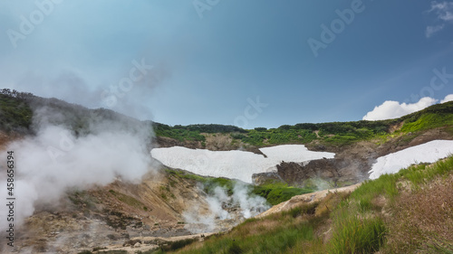 Clouds of vapor from fumaroles rise into the blue sky. On the mountain slopes, among the green vegetation, there is melted snow. Blue sky. The valley of hot springs. Kamchatka
