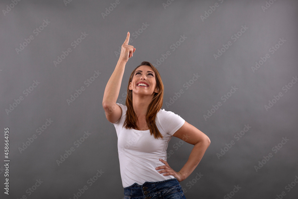Cheerful brunette model dressed in casual clothes smiles and points upwards with finger isolated on gray background
