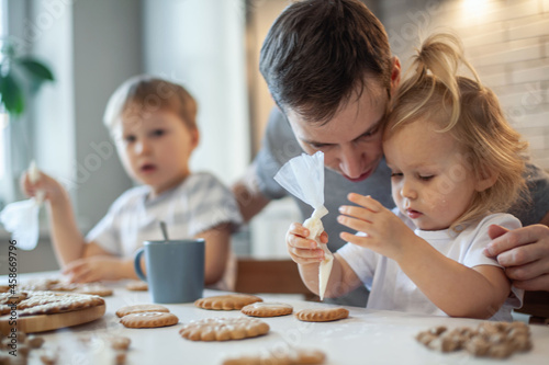 Dad and children decorate Christmas gingerbread at home. A boy and a girl paint with cornets with sugar icing on cookies. New Year's decor, branches of a Christmas tree.