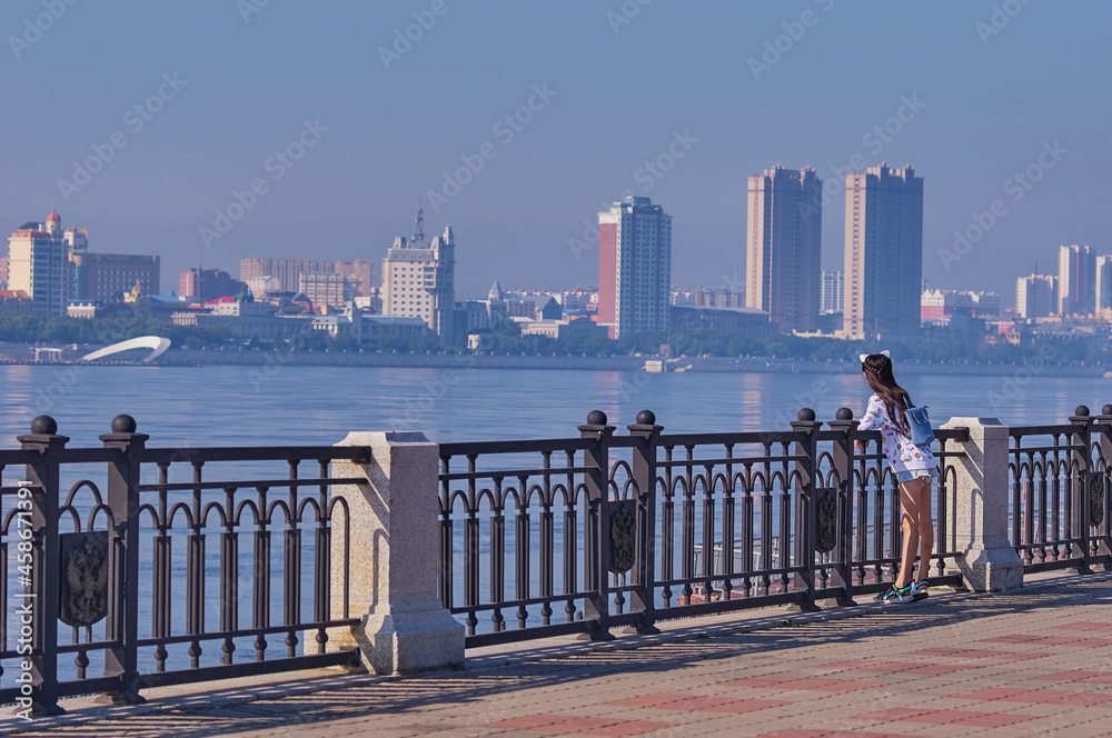 View from the embankment of the Amur river in the city of Blagoveshchensk, Russia. A young girl admires the view of the city of Heihe, China. Focus on the foreground.
