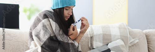 Fotografie Young woman wrapped in warm blanket sits on sofa with thermometer
