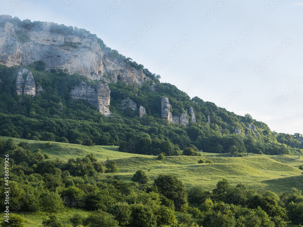 A mighty mountain with sheer cliffs rises in the background of small green trees. Above it is a blue sky. Morning landscape. A mighty mountain with sheer cliffs rises in the background of small green trees. Above it is a blue sky. Morning landscape.