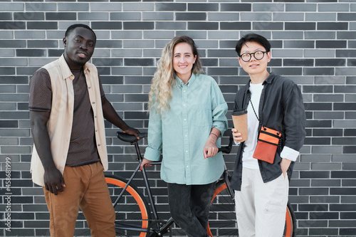 Horizontal medium long portrait of young ethnically diverse friends wearing casual outfits standing together against gray brick building looking at camera