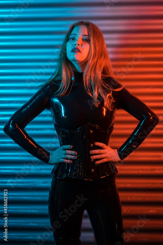 Red-haired girl with latex, a black top and tight pants, on a metallic background illuminated with red and blue LEDs, posed looking at the camera photo