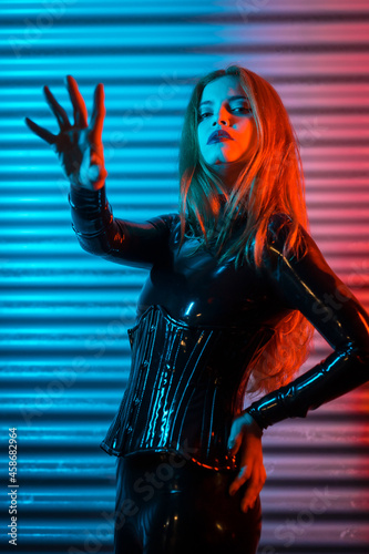 Red-haired girl wearing latex, a black top and tight pants, on a metallic background illuminated with red and blue LEDs, making the stop gesture photo