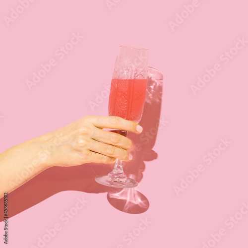 Celebrity concept. Girl hand holding antique crystal glass with red drink in it against baby pink background