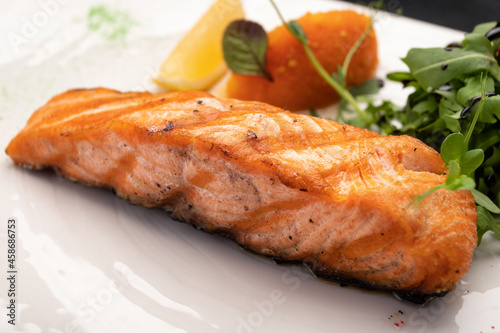 Fried salmon steak with herbs and lemon on a white plate on a dark background
