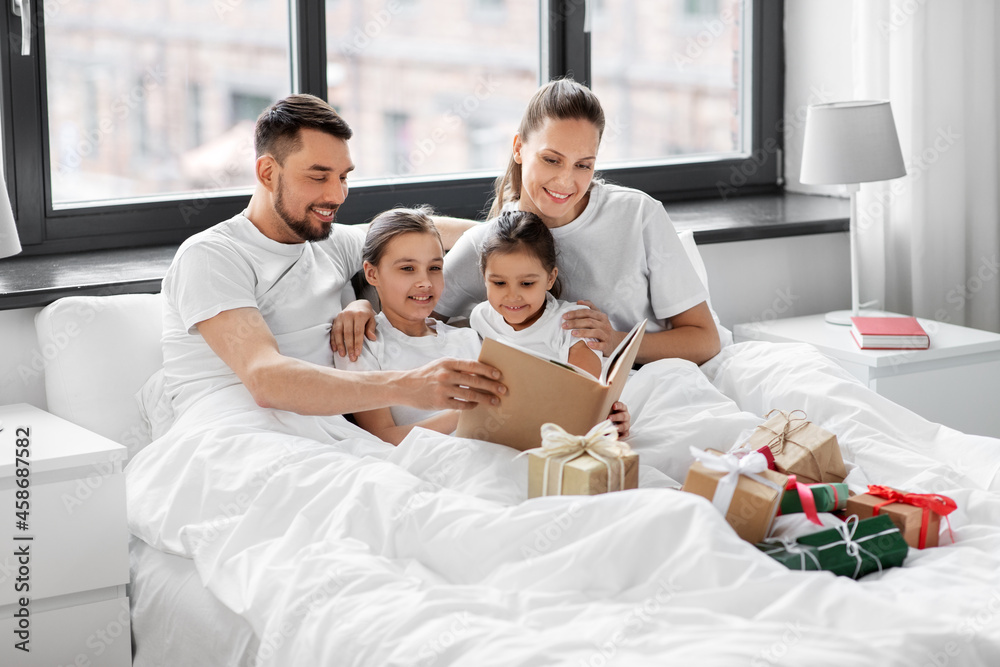 family, winter holidays and people concept - happy mother, father and two daughters in pajamas with christmas gifts reading book in bed at home