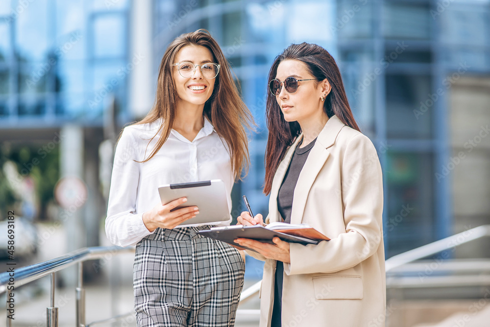 Two young business lady with tablet and notebook walking outdoors near modern business center. Two young business lady with tablet and notebook walking outdoors near modern business center.