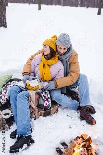 Man hugging girlfriend with cup of coffee near bonfire in winter park