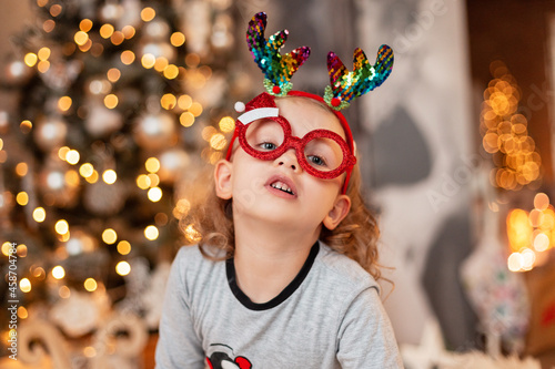 Funny little girl with fun red Santa glasses and deer horns in fashionable pajamas against the backdrop of Christmas decorations at home