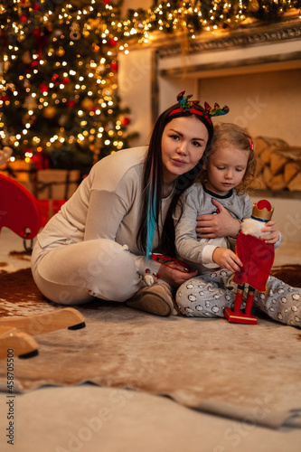 Happy beautiful mother with her daughter child are sitting near the fireplace and Christmas decorations and playing with a toy together. Winter family holidays