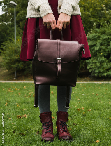 Front view of female legs in grey tights and skirt. Stylish burgundy shoes. Leather bag in the arms. Green grass. Autumn fashion.