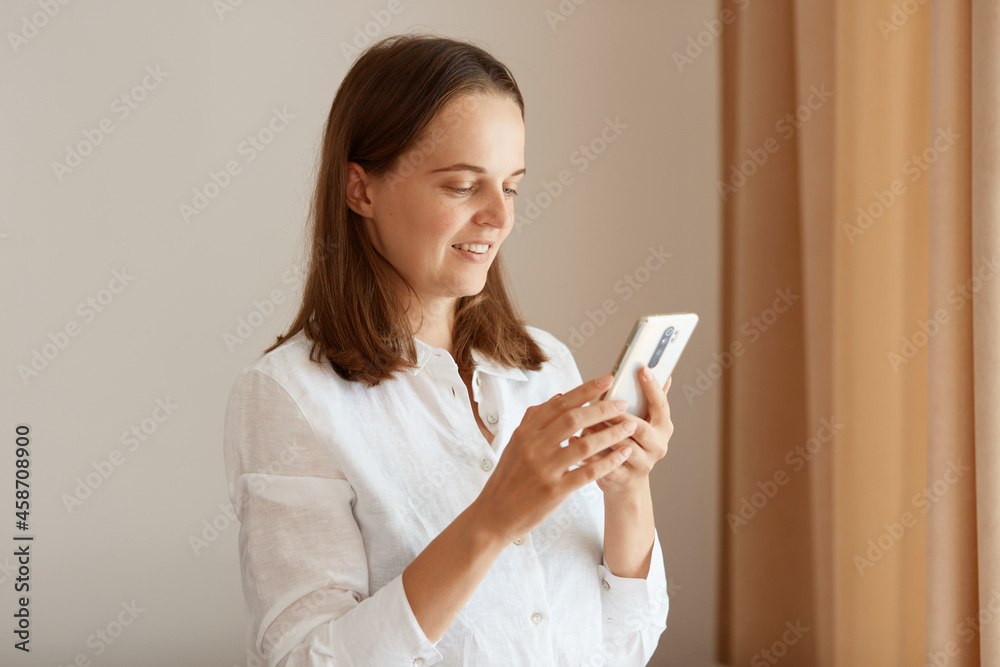 Side view portrait of positive woman with dark hair wearing white cotton shirt standing with cell phone in hands near window in living room at home, browsing internet. Side view portrait of positive woman with dark hair wearing white cotton shirt standing with cell phone in hands near window in living room at home, browsing internet.