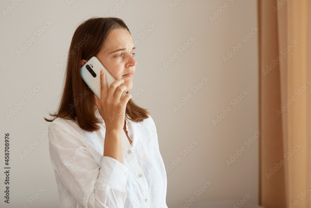 Side view of positive woman wearing white cotton shirt standing near window with beige cottons and talking phone, looking away, having calm facial expression. Side view of positive woman wearing white cotton shirt standing near window with beige cottons and talking phone, looking away, having calm facial expression.
