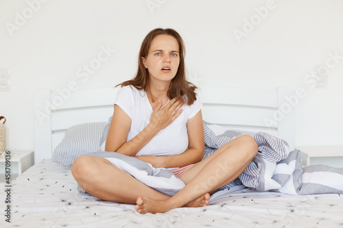 Indoor shot of sick woman wearing white casual t shirt, sitting on bed with crossed legs, touching her chest, suffering from heart pain, looking at camera with frowning face.