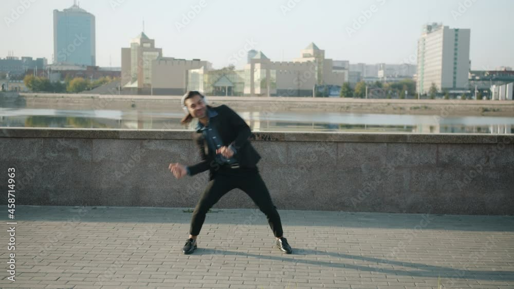 Joyful guy wearing headphones is dancing outdoors at river embankment enjoying music and having fun on summer day. Street dance and emotions concept. Joyful guy wearing headphones is dancing outdoors at river embankment enjoying music and having fun on summer day. Street dance and emotions concept.