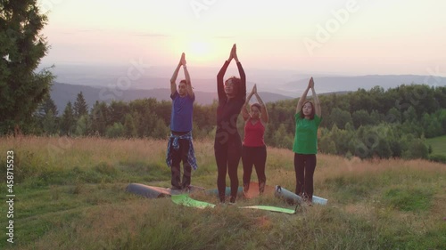 Serene diverse multiethnic sporty fit people meditating, standing in sun salutation posture, practicing spiritual healing and release tension while doing group yoga class on mountain top at daybreak.