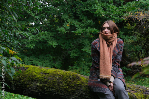 Young red-haired beautiful girl sits in the forest on a fallen tree overgrown with moss in autumn clothes. High quality photo