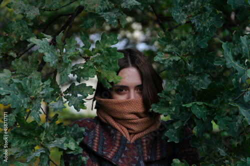beautiful young woman with red hair looks out from behind the leaves of a tree in the park in autumn. High quality photo