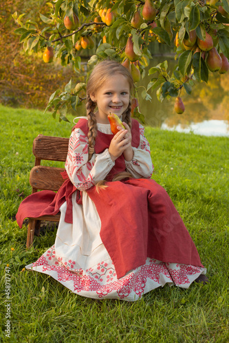 A BLONDE GIRL IN A SLAVIC COSTUME IS SITTING ON A BENCH NEAR A TREE WITH PEARS LAKE BRANCHES AUTUMN