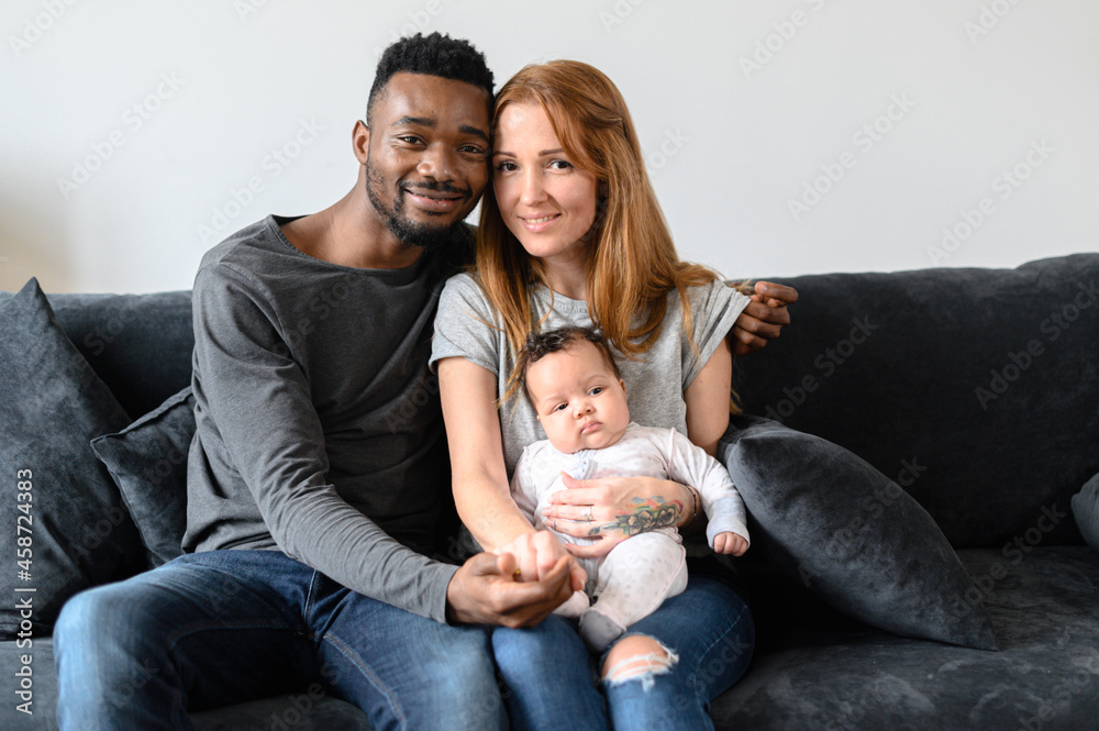 Portrait of happy mixed-race family of three African-American husband, caucasian wife and a cute biracial daughter sitting on the couch indoors and looking at the camera Portrait of happy mixed-race family of three African-American husband, caucasian wife and a cute biracial daughter sitting on the couch indoors and looking at the camera