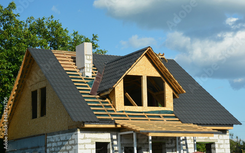 Metal roofing construction. Metal roof tiles installation on a gable roof with attic windows and chimney of a house under construction.