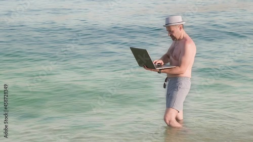 Male in the striped swimming trunks and hat working remotely at the vacation from the sea and ocean. Man workaholic stands in the water with the laptop and typing report or doing distant help desk.