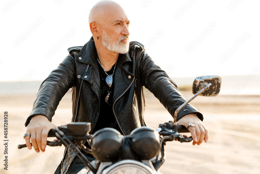 Bold senior man wearing leather jacket riding motorcycle outdoors
