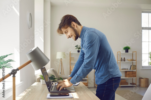 Happy young businessman or freelancer writing text on notebook while working at home office. Side view of smiling man standing at his desk searching for information on internet or writing email.