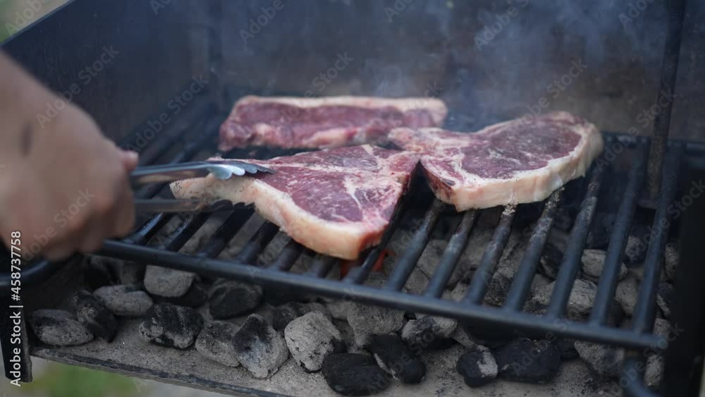 Tongs flip raw steaks grilling over hot smoking coals.