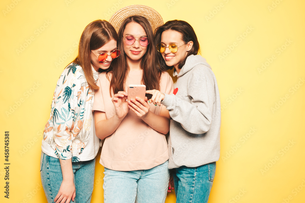 Three young beautiful smiling hipster female in trendy clothes.Sexy carefree women posing near yellow wall in studio.Positive models looking at smartphone screen. Using cellphone apps Three young beautiful smiling hipster female in trendy clothes.Sexy carefree women posing near yellow wall in studio.Positive models looking at smartphone screen. Using cellphone apps