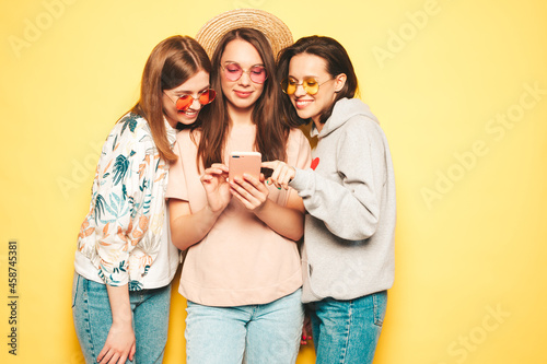 Three young beautiful smiling hipster female in trendy clothes.Sexy carefree women posing near yellow wall in studio.Positive models looking at smartphone screen. Using cellphone apps