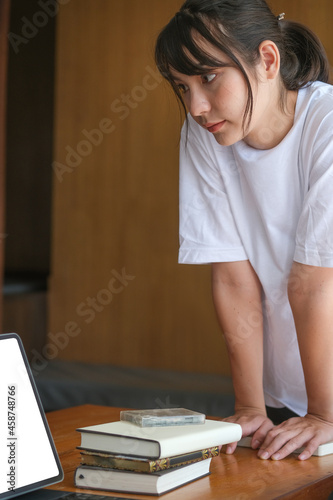 Photo of a cute student girl standing with a hand leaning on the table surrounded by a digital tablet and stack of books.