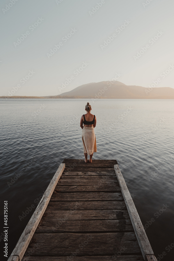 Young woman standing at the end of a jetty looking over a lake with a mountain in the background. Young woman standing at the end of a jetty looking over a lake with a mountain in the background.
