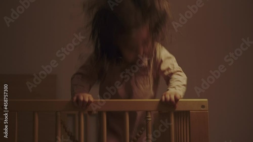 Little energetic happy girl jumping in a crib, does not want to sleep, plays. There is artificial light from a lamp in the children's room. Dust.