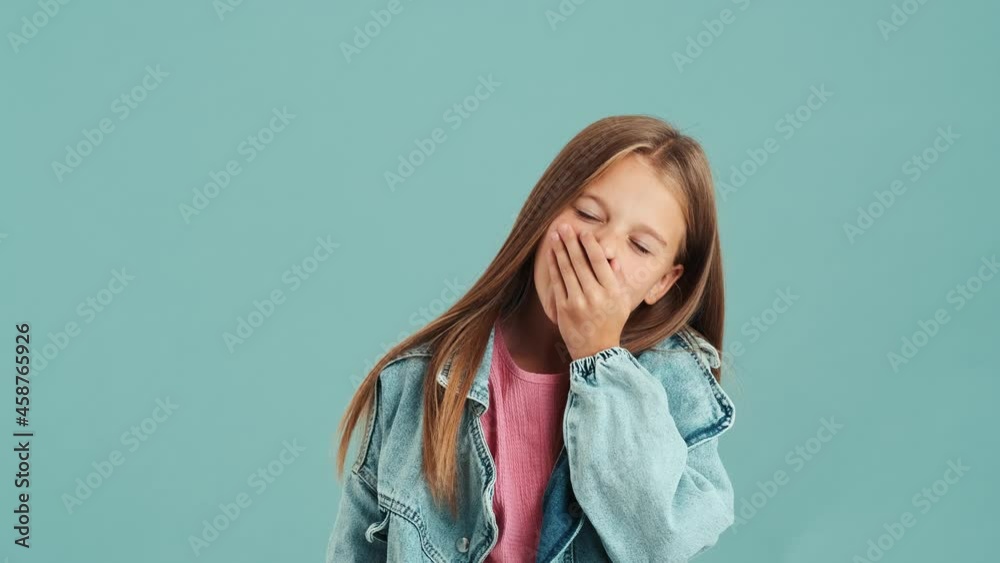 A sleepy bored small girl wearing jeans jacket yawning standing isolated over a blue wall in the studio A sleepy bored small girl wearing jeans jacket yawning standing isolated over a blue wall in the studio