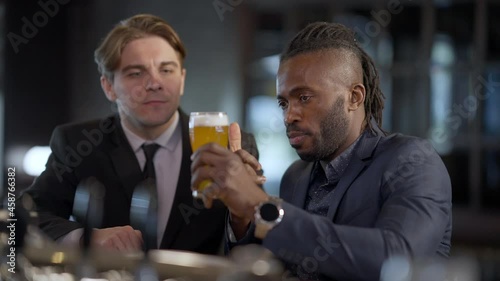 African American beer connoisseur talking with Caucasian friend explaining pale lager features pointing at glass. Portrait of concentrated men in formal suits resting in bar after work indoors photo