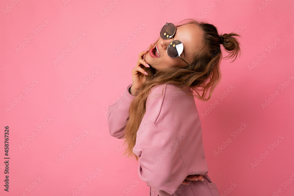 Shot of beautiful sexy happy young dark blonde curly woman isolated over pink background wall wearing casual pink sport clothes and stylish sunglasses looking at camera Shot of beautiful sexy happy young dark blonde curly woman isolated over pink background wall wearing casual pink sport clothes and stylish sunglasses looking at camera