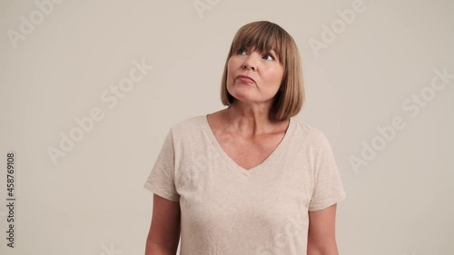 An unknowing mature woman with bob hairstyle spreading her hands to the sides on gray-beige background in studio photo