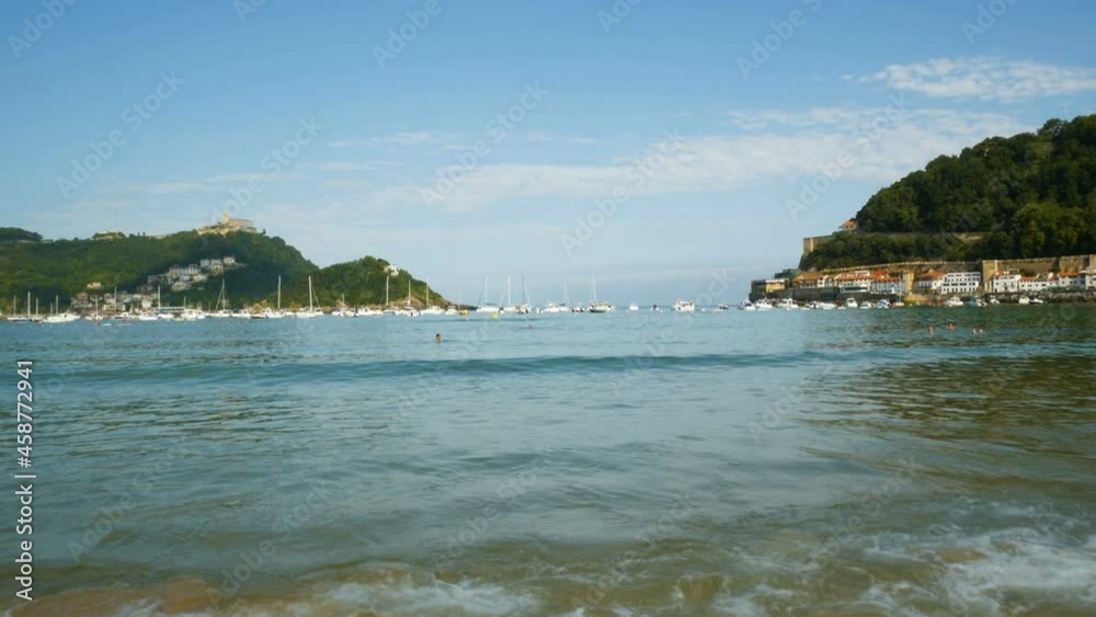 The calm ocean waters of the peaceful bay of San Sebastian, Spain. Little waves in the foreground. Small harbor and blue sky on the background. The calm ocean waters of the peaceful bay of San Sebastian, Spain. Little waves in the foreground. Small harbor and blue sky on the background.