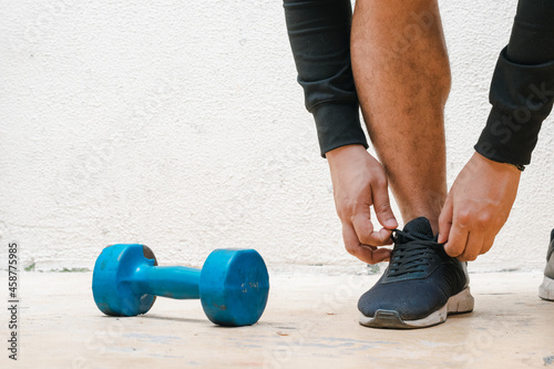 man tying his shoe standing next to blue dumbbell, before exercising. exercise at home, white background. fitness concept. health concept, space for advertising text