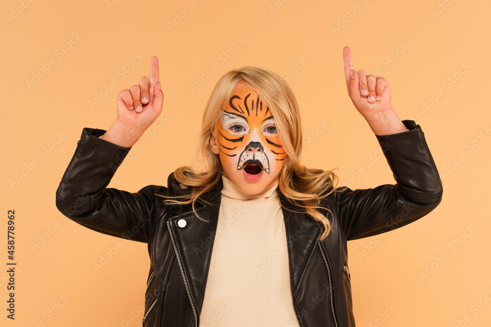 thrilled kid in tiger makeup pointing up with fingers while looking at camera isolated on beige thrilled kid in tiger makeup pointing up with fingers while looking at camera isolated on beige