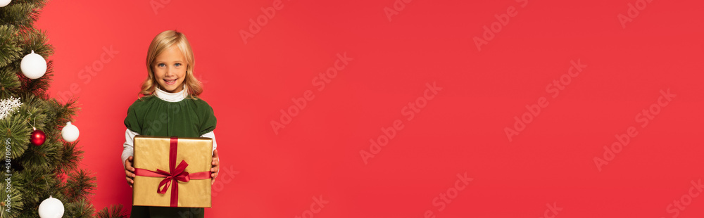 happy girl holding present near decorated christmas tree isolated on red, banner happy girl holding present near decorated christmas tree isolated on red, banner