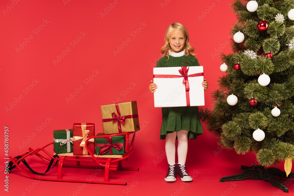 happy girl holding present near christmas tree and sled with gift boxes on red happy girl holding present near christmas tree and sled with gift boxes on red