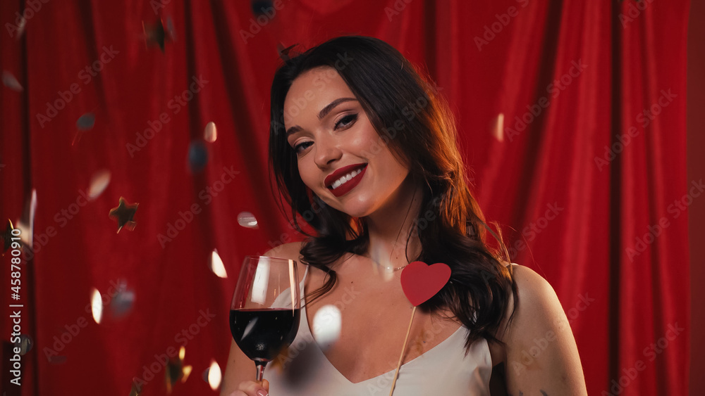 happy woman holding glass of wine near falling confetti on red. happy woman holding glass of wine near falling confetti on red.