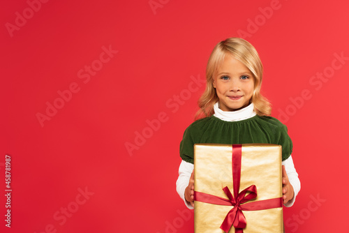 smiling child holding present in golden wrapping paper while looking at camera isolated on red