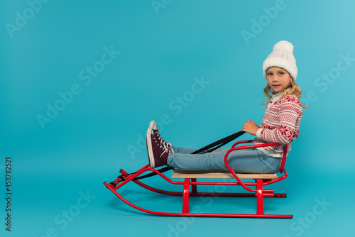 kid in knitted hat and sweater sitting in sled and looking at camera on blue