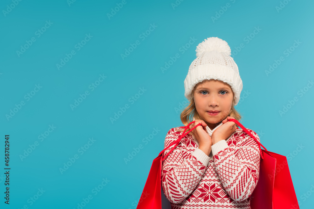 girl in knitted hat and jumper with ornament holding purchases isolated on blue girl in knitted hat and jumper with ornament holding purchases isolated on blue